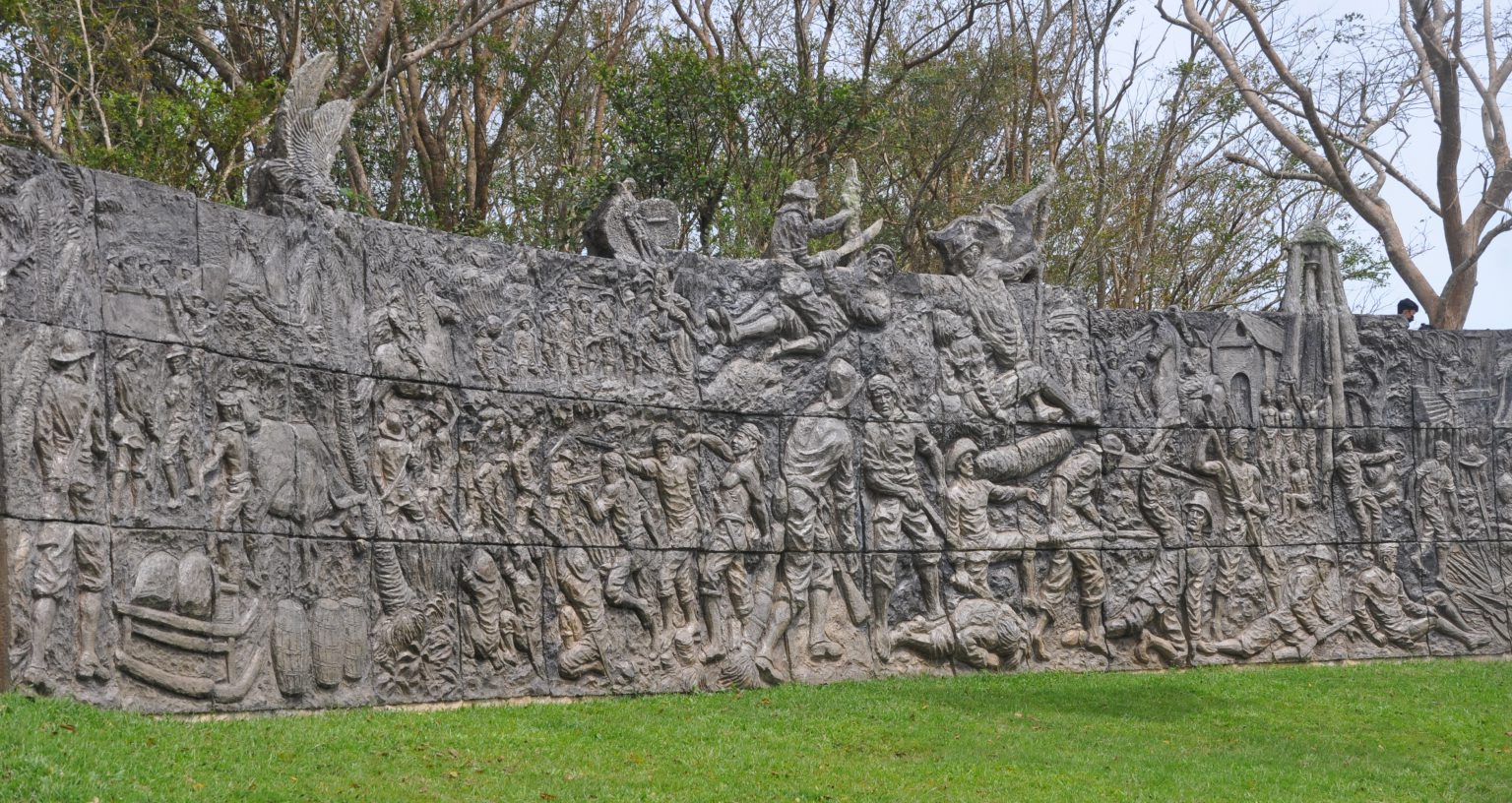 Stone relief mural at Pulang Lupa Historical Park in Torrijos, Marinduque, showing Filipino patriots ambushing American soldiers during the Battle of Pulang Lupa in 1900, with detailed carvings of combat scenes, surrender, and the island’s terrain, set against a grassy foreground and tree-lined background